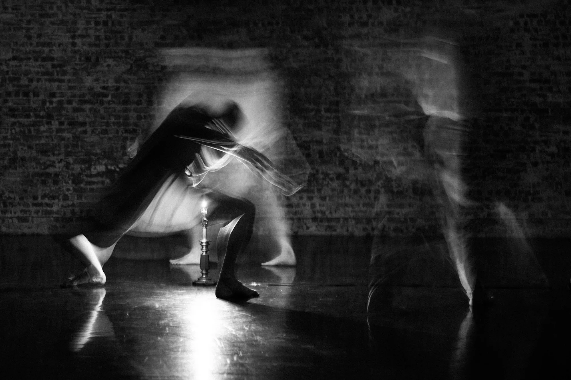 Black-and-white photograph of a dancer in motion beside a small floor lamp against a brick wall.