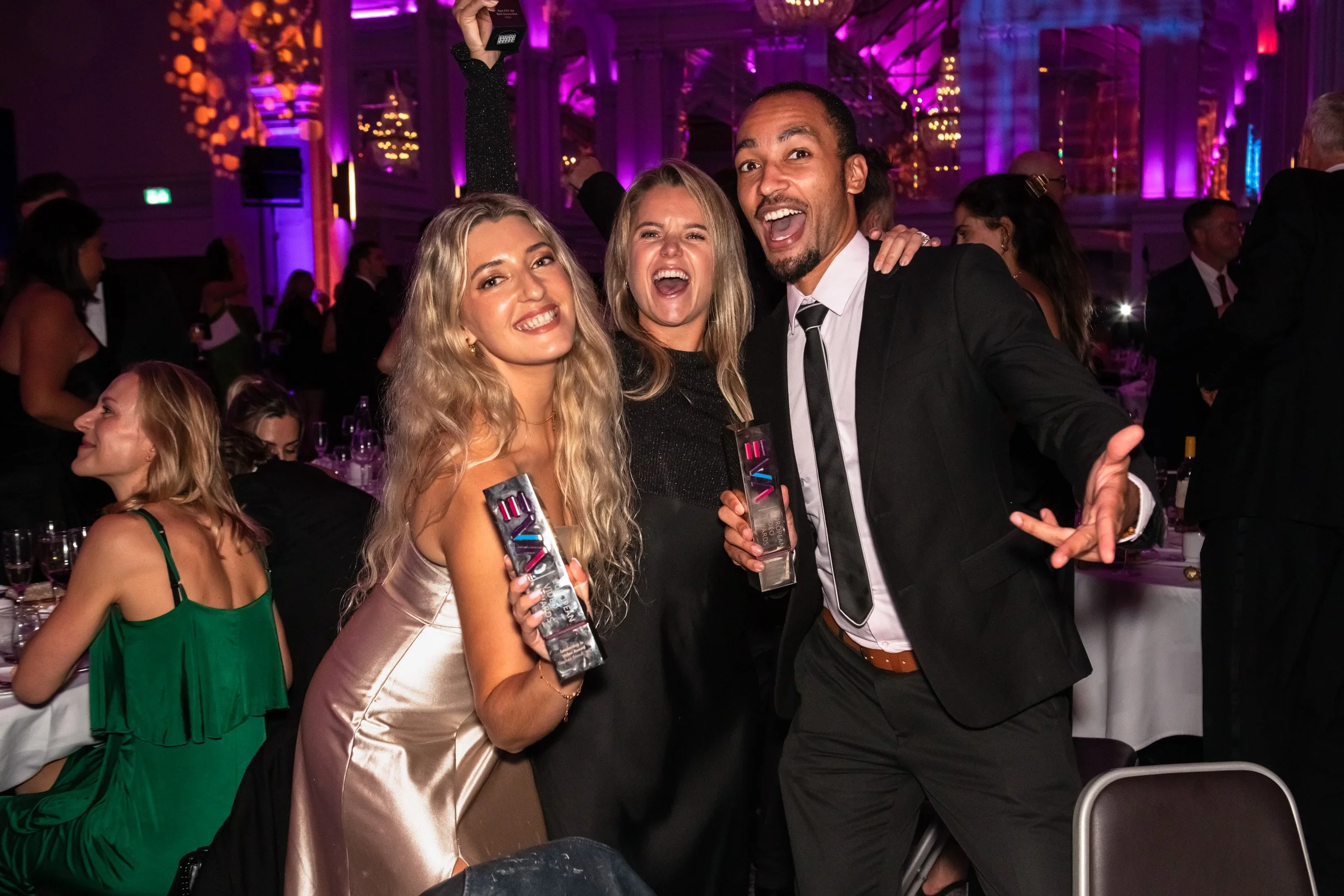 Three award winners smiling and celebrating while holding trophies at an awards ceremony with purple lighting.