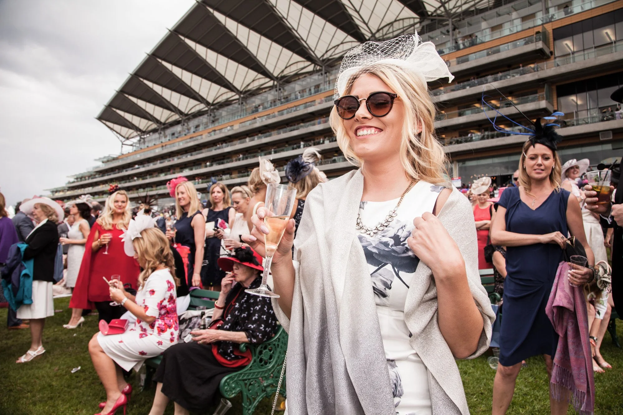 A guest in sunglasses and a cream jacket smiling among the crowd at Royal Ascot.