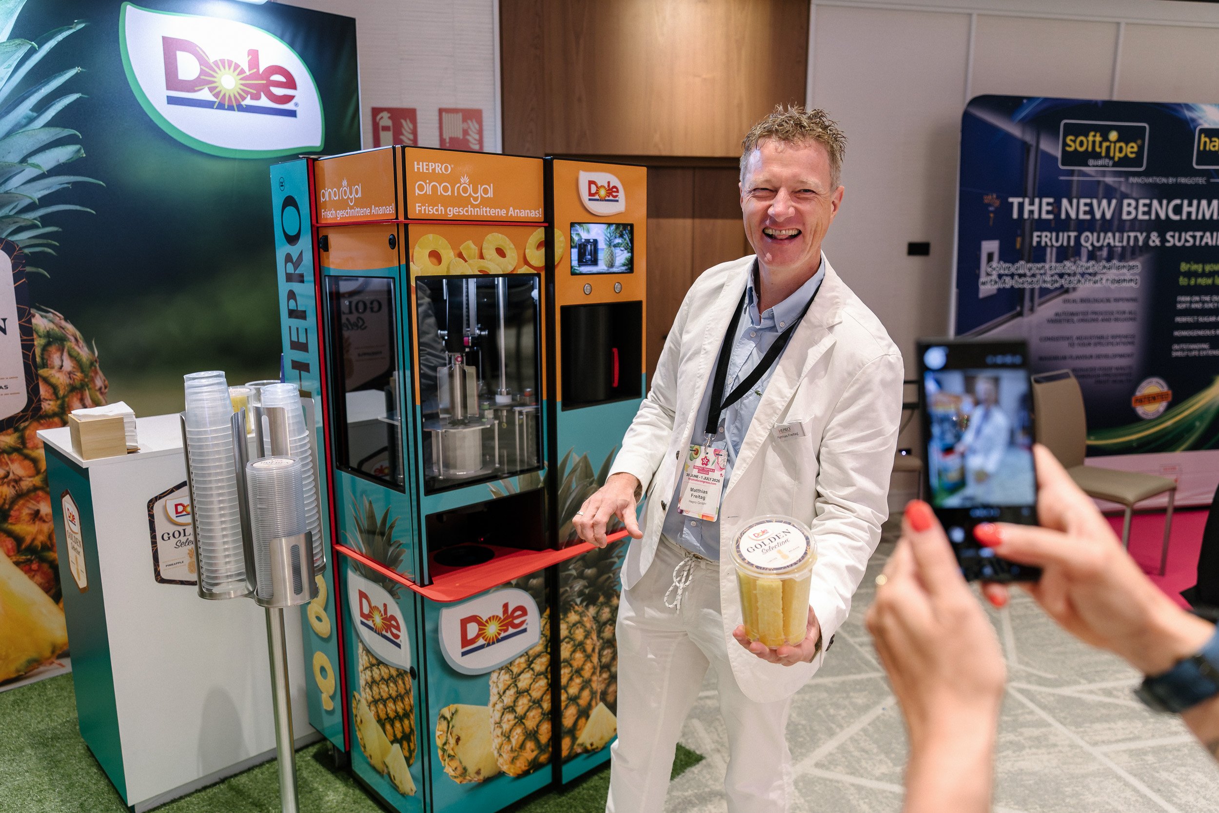Smiling Dole representative demonstrating a pineapple product at a trade show while an attendee takes a photograph with a smartphone.