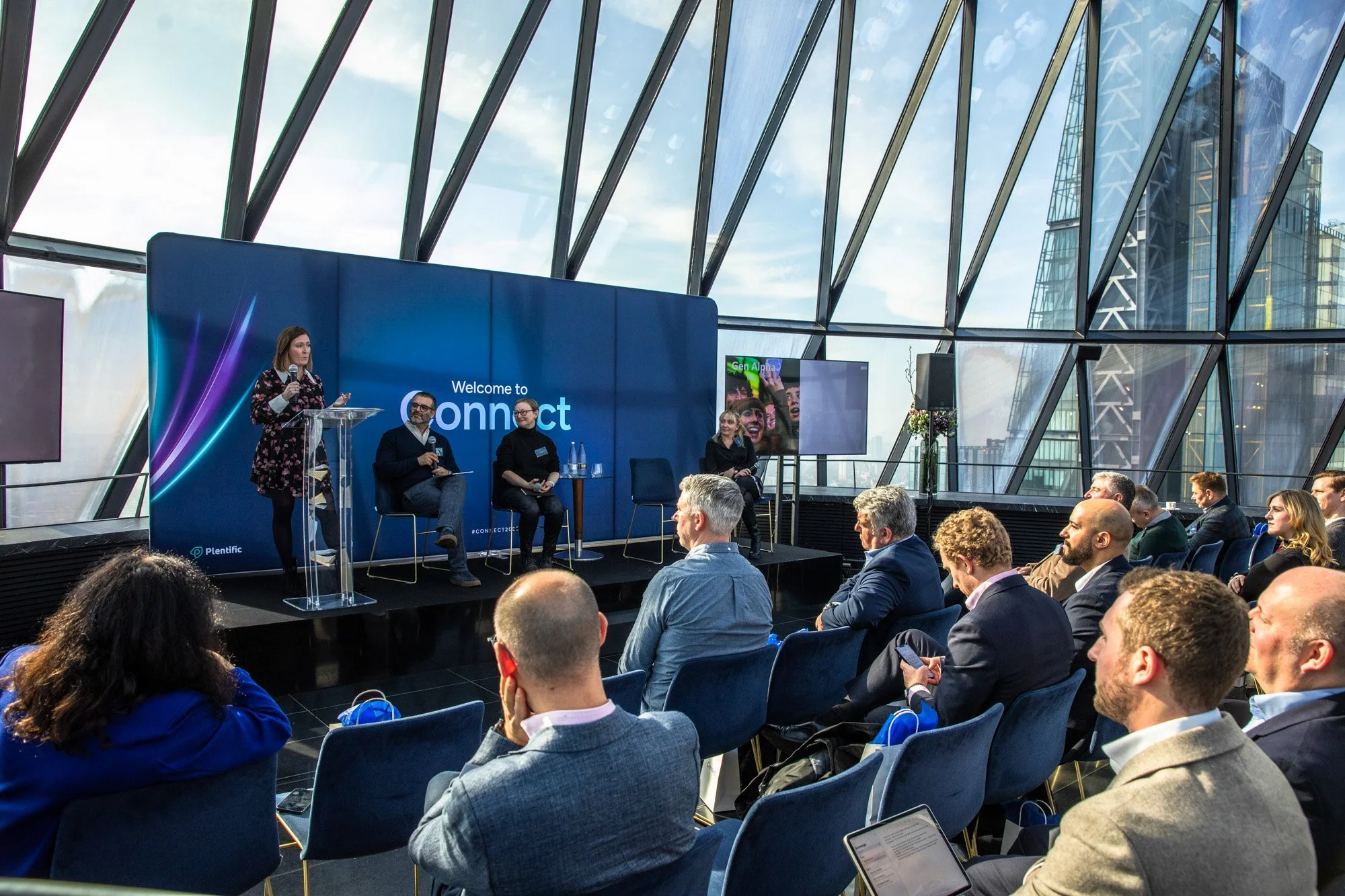 Corporate conference at The Gherkin in London, with a speaker presenting on stage to an audience during the Connect+ business event.