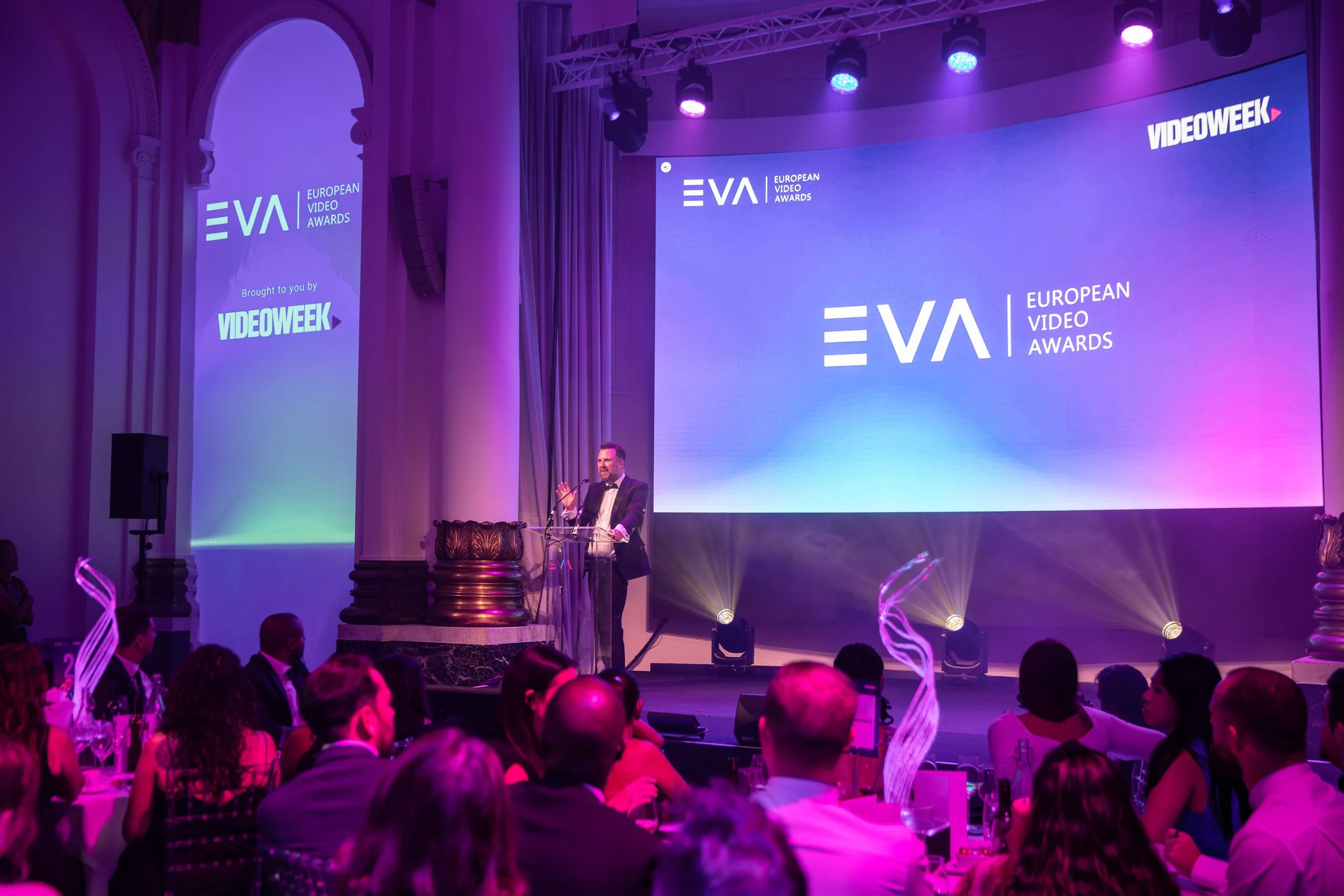 Presenter speaking on stage at the European Video Awards, with purple and blue lighting, large branded screens and guests seated at tables.