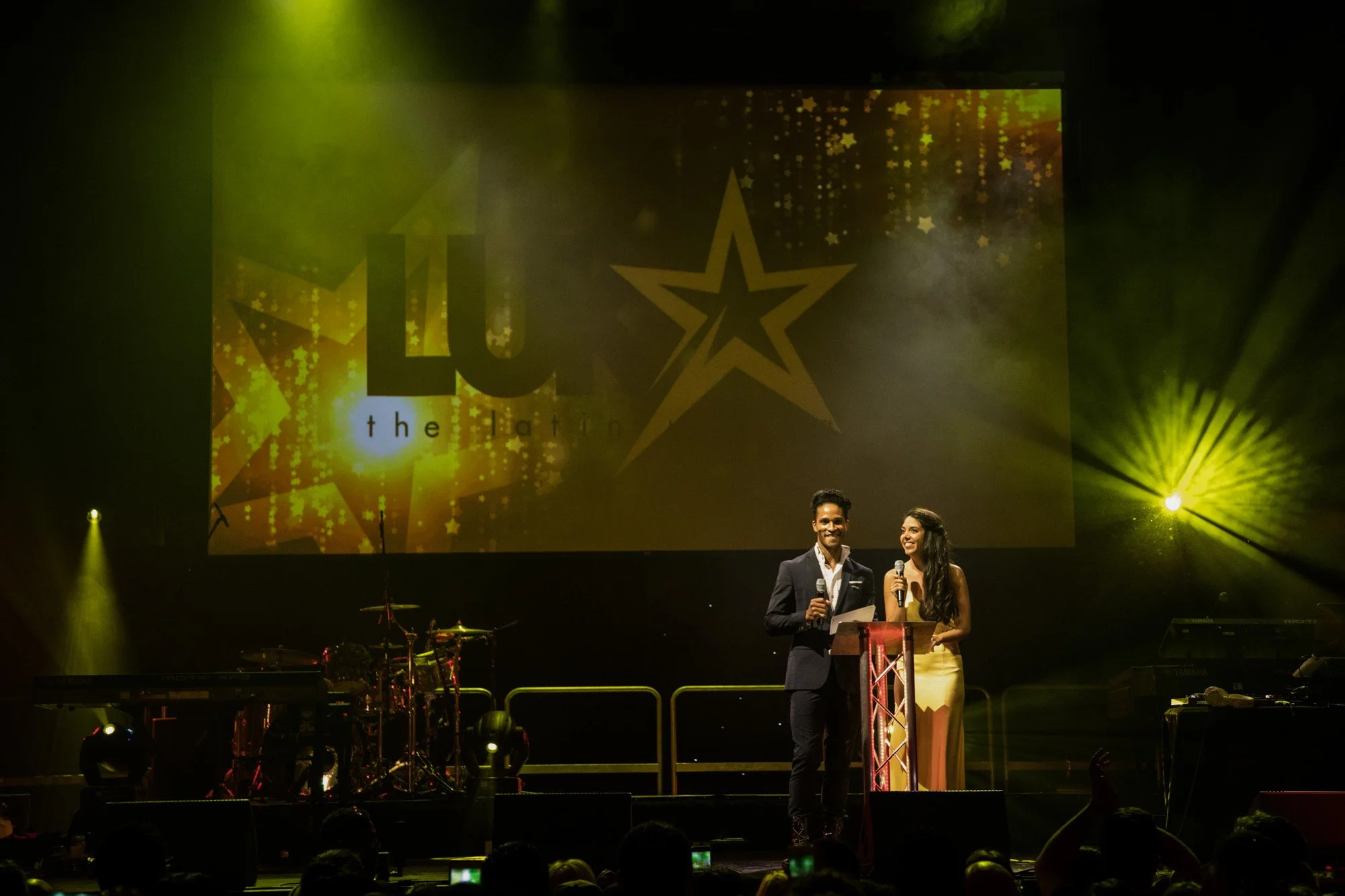 Two presenters on the LUKAS Awards stage under yellow spotlights.