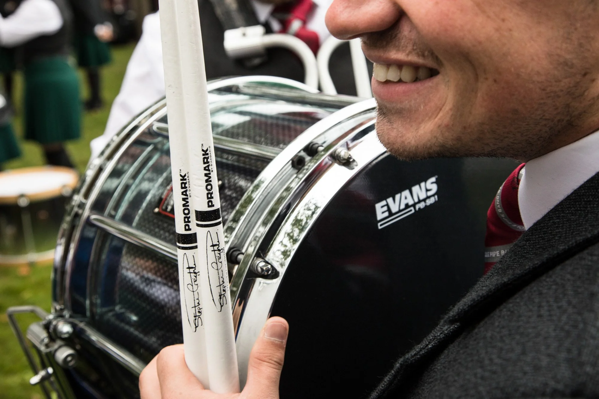 A smiling drummer holding drumsticks beside a bass drum at a pipe-band event.
