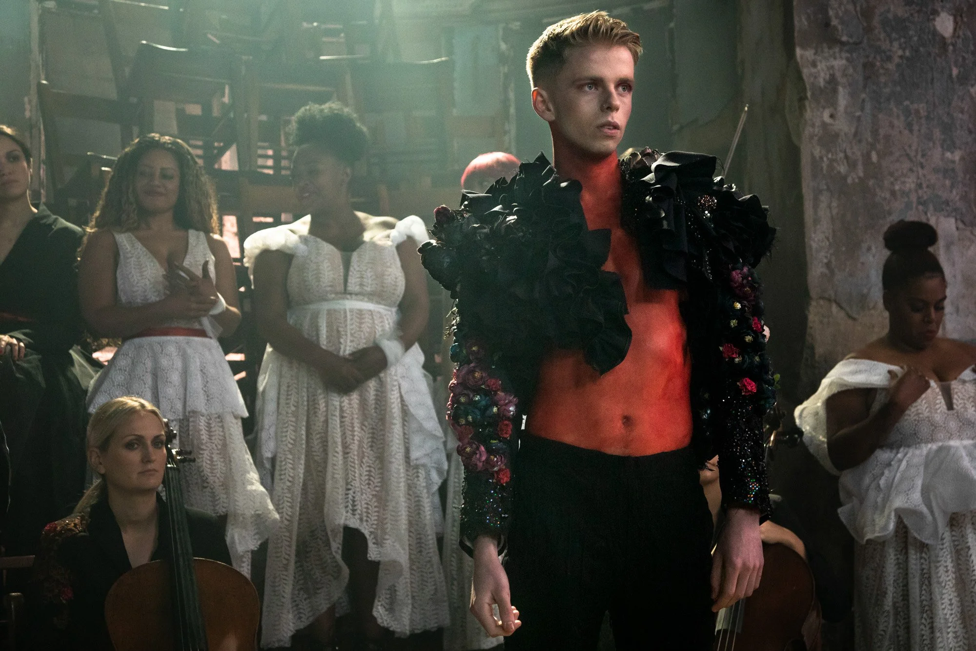 A performer in a floral jacket standing in front of an ensemble wearing white lace costumes.