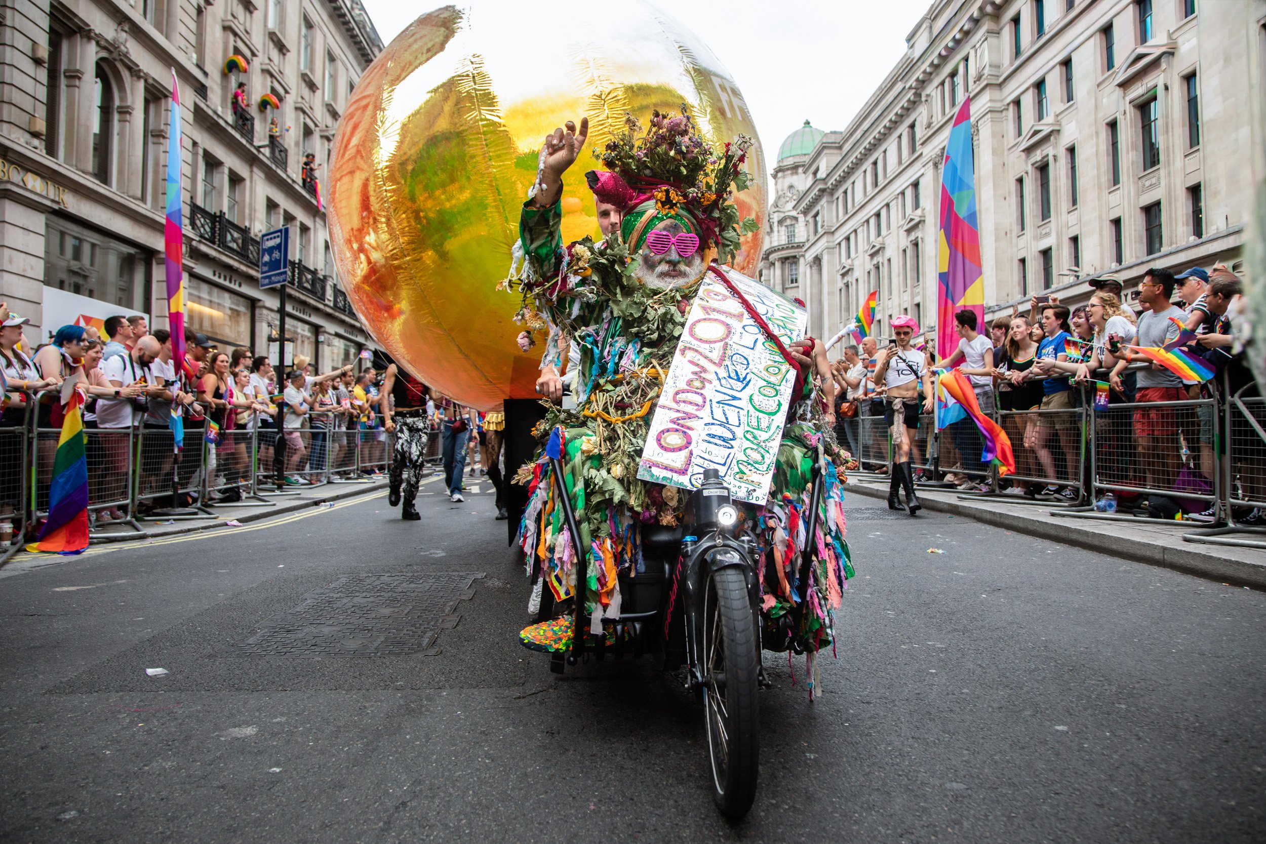 Colourfully dressed participant riding a decorated bicycle in front of a giant golden balloon during the London Pride parade, with crowds cheering on either side.