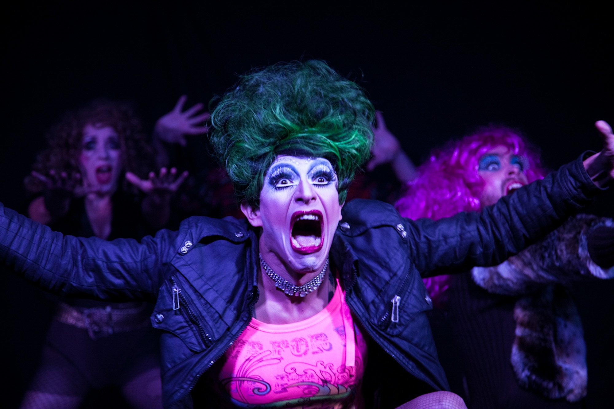 A green-haired performer in colourful makeup and a leather jacket on stage with an ensemble.