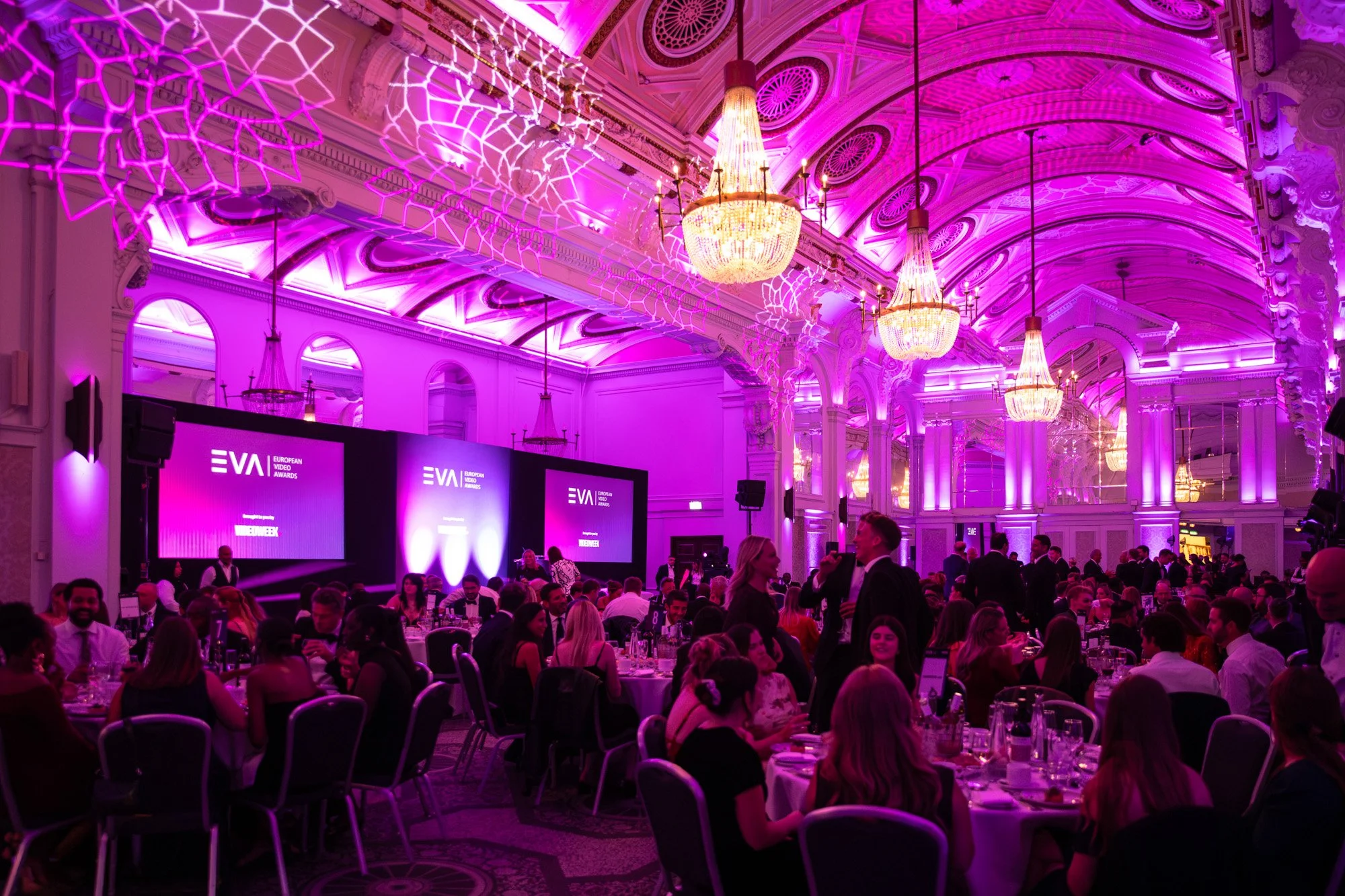 Guests seated beneath chandeliers in a grand ballroom lit in purple for an awards dinner.