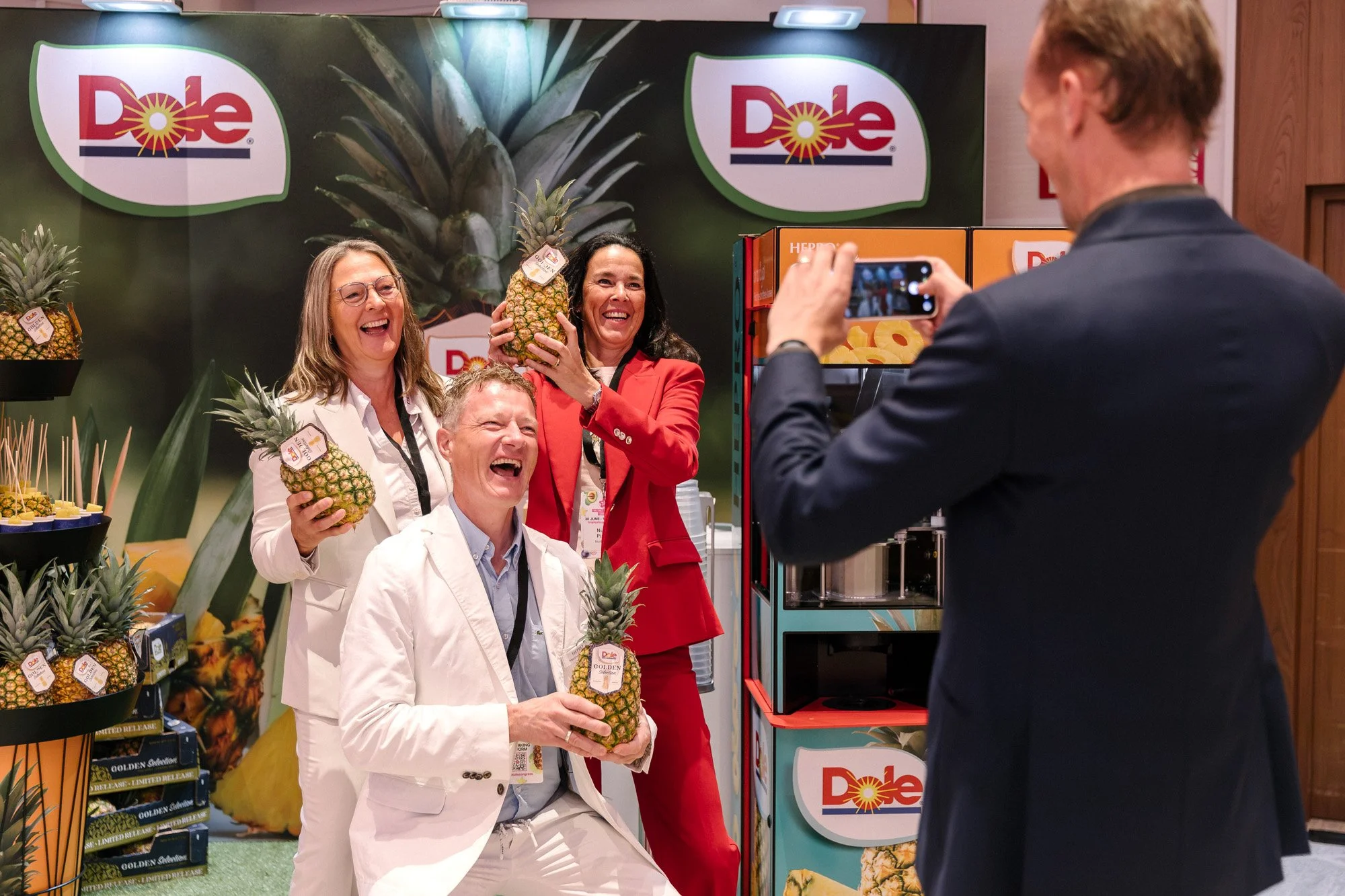 Visitors posing with pineapples at the Dole exhibition stand while another guest takes a photograph.