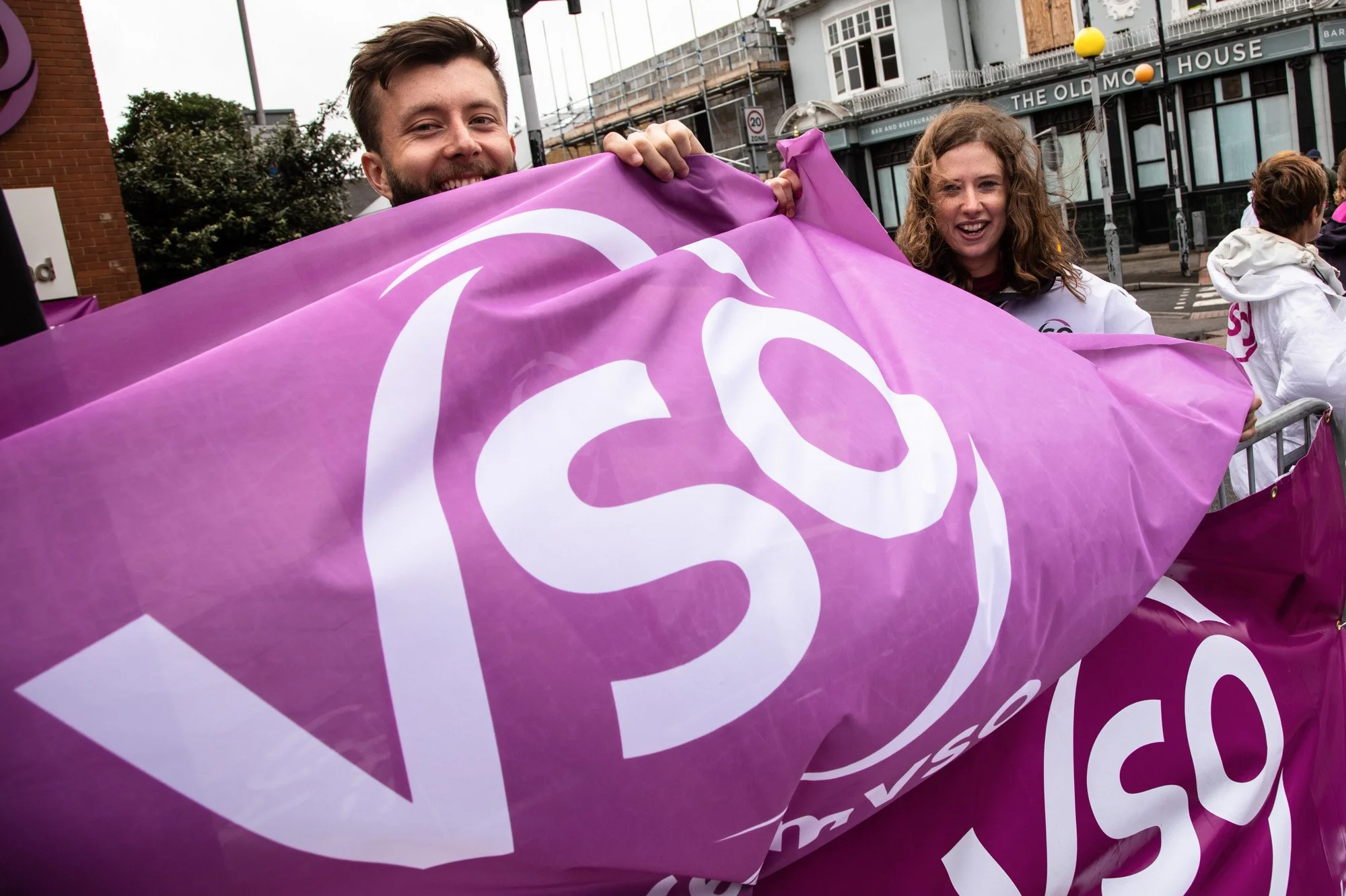 Two smiling VSO supporters holding a large purple banner at RideLondon.