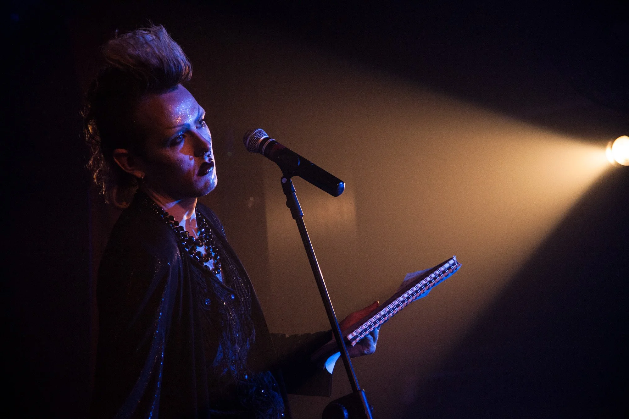 A performer reading from a notebook at a microphone under a spotlight.