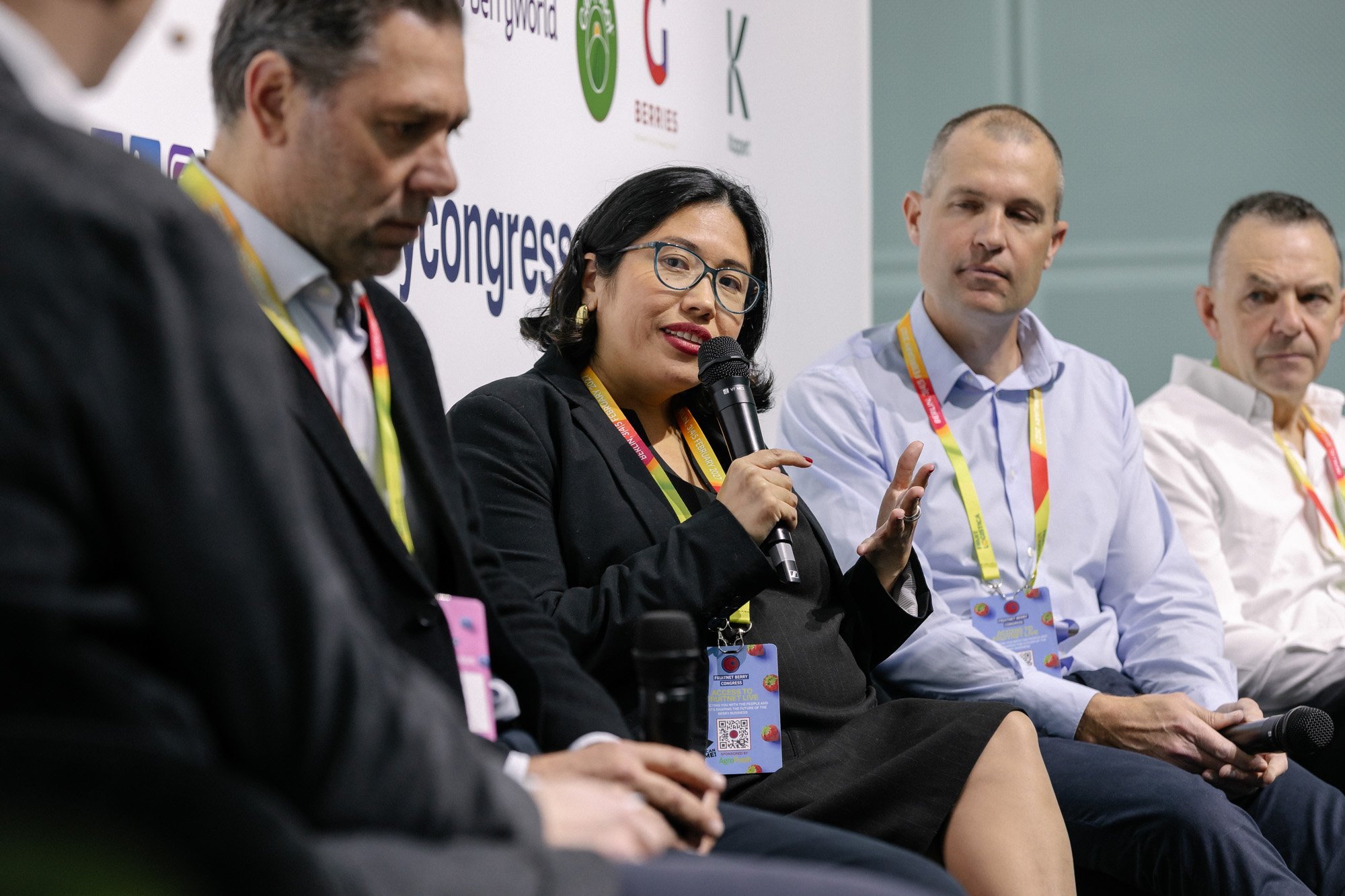 A woman in glasses speaking into a microphone beside other attendees during a conference discussion.