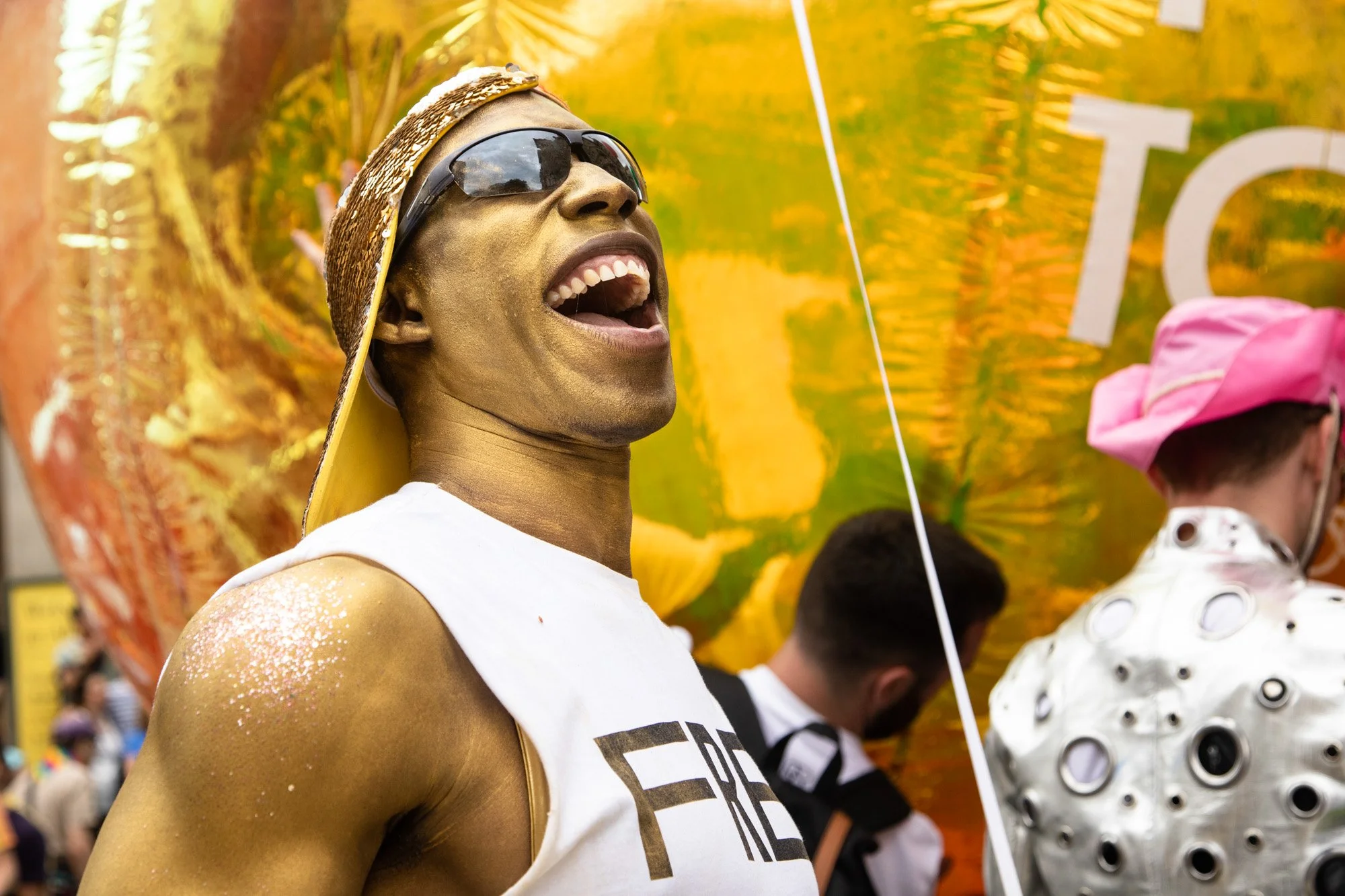 A smiling Pride participant in sunglasses and gold body paint beside a gold balloon.