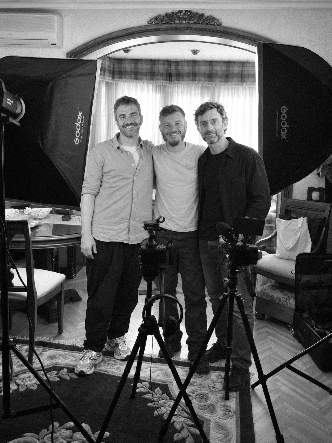 Black-and-white portrait of three MVV Media collaborators standing together on a filming set, surrounded by cameras, tripods and studio lights.
