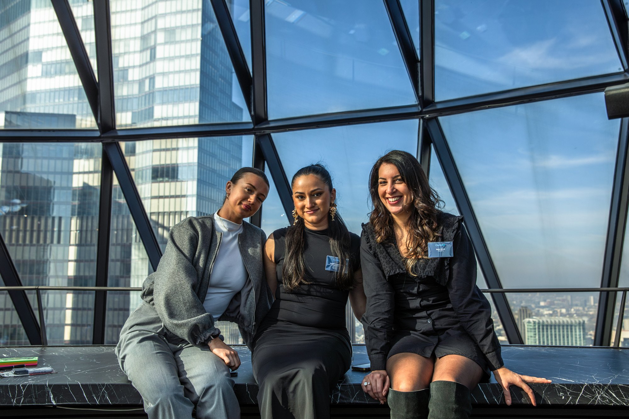 Three event attendees seated together in front of large windows overlooking the city.