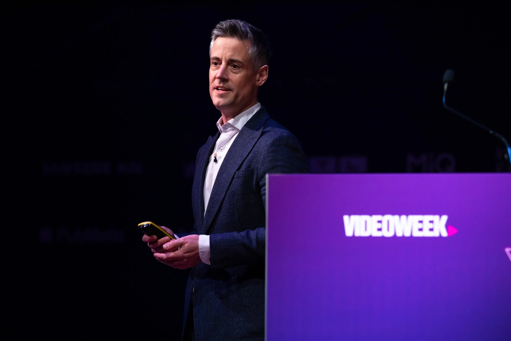 A speaker in a dark suit presenting beside a purple VideoWeek lectern.