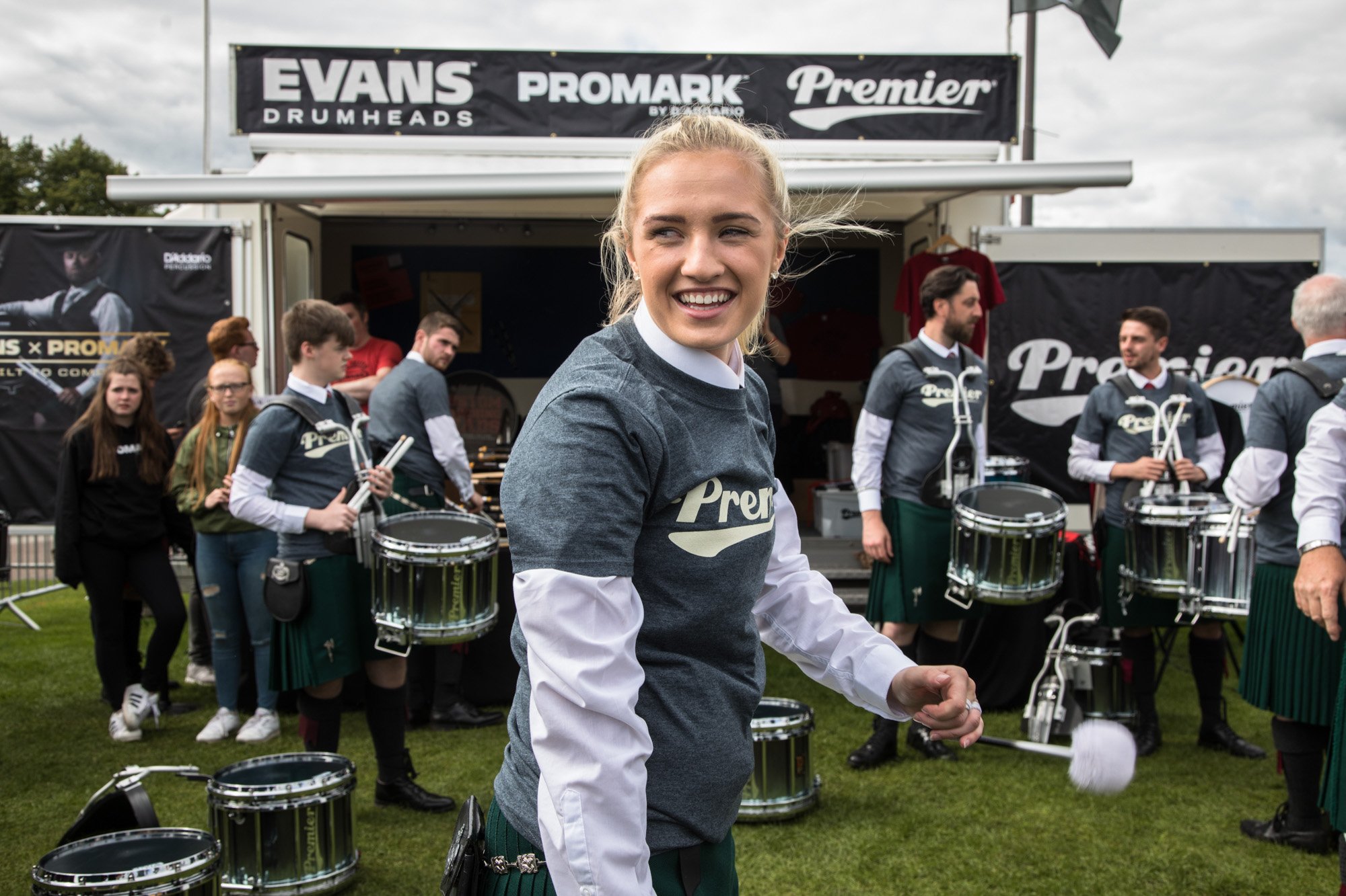 A smiling drummer holding mallets beside a group of musicians at a pipe-band event.