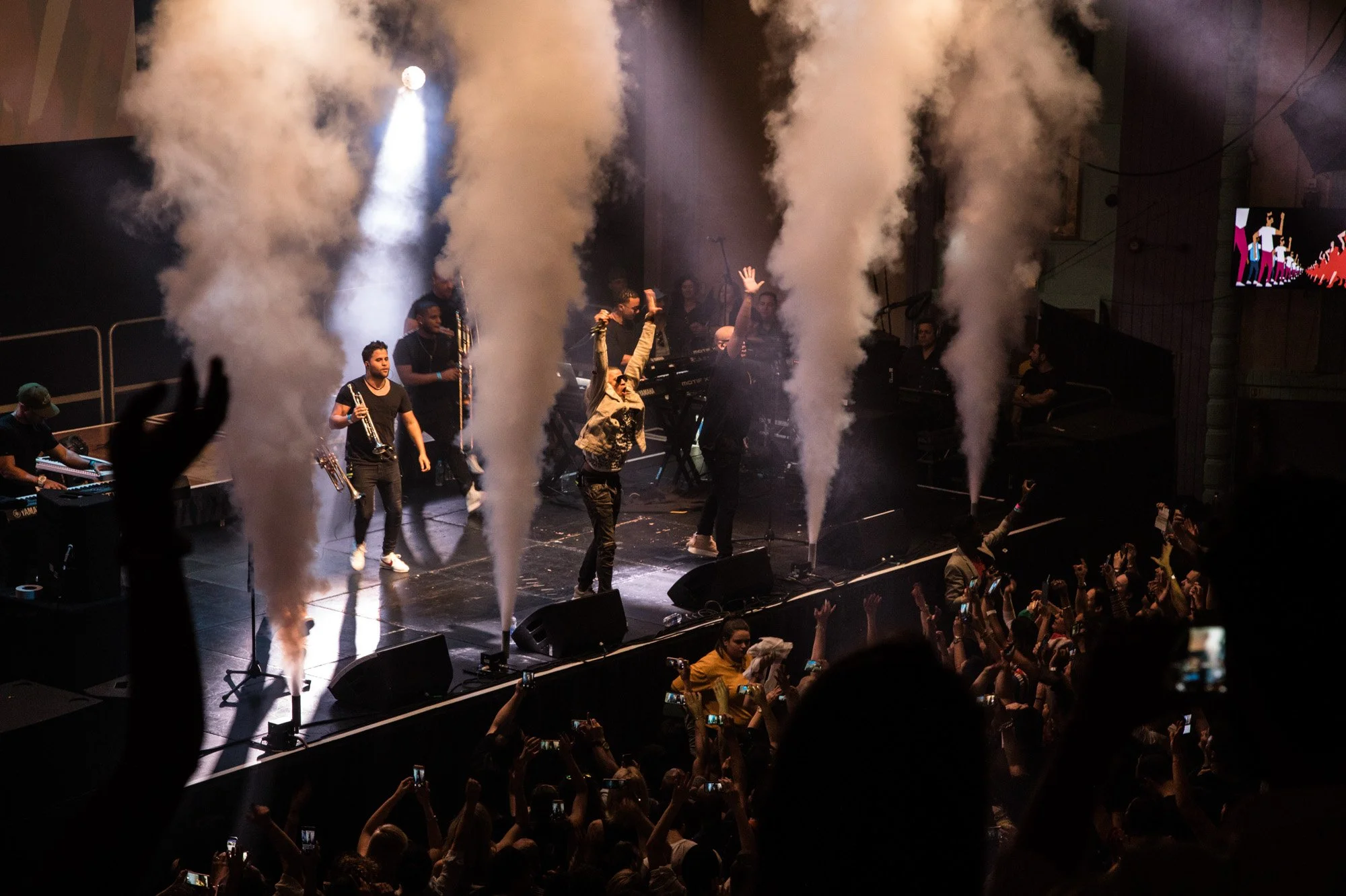 Musicians performing on stage amid columns of smoke, with fans watching from the crowd.