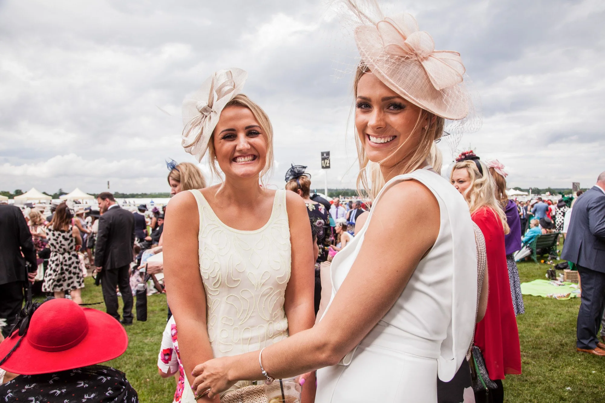 Two guests in pale dresses and decorative hats smiling together at Royal Ascot.