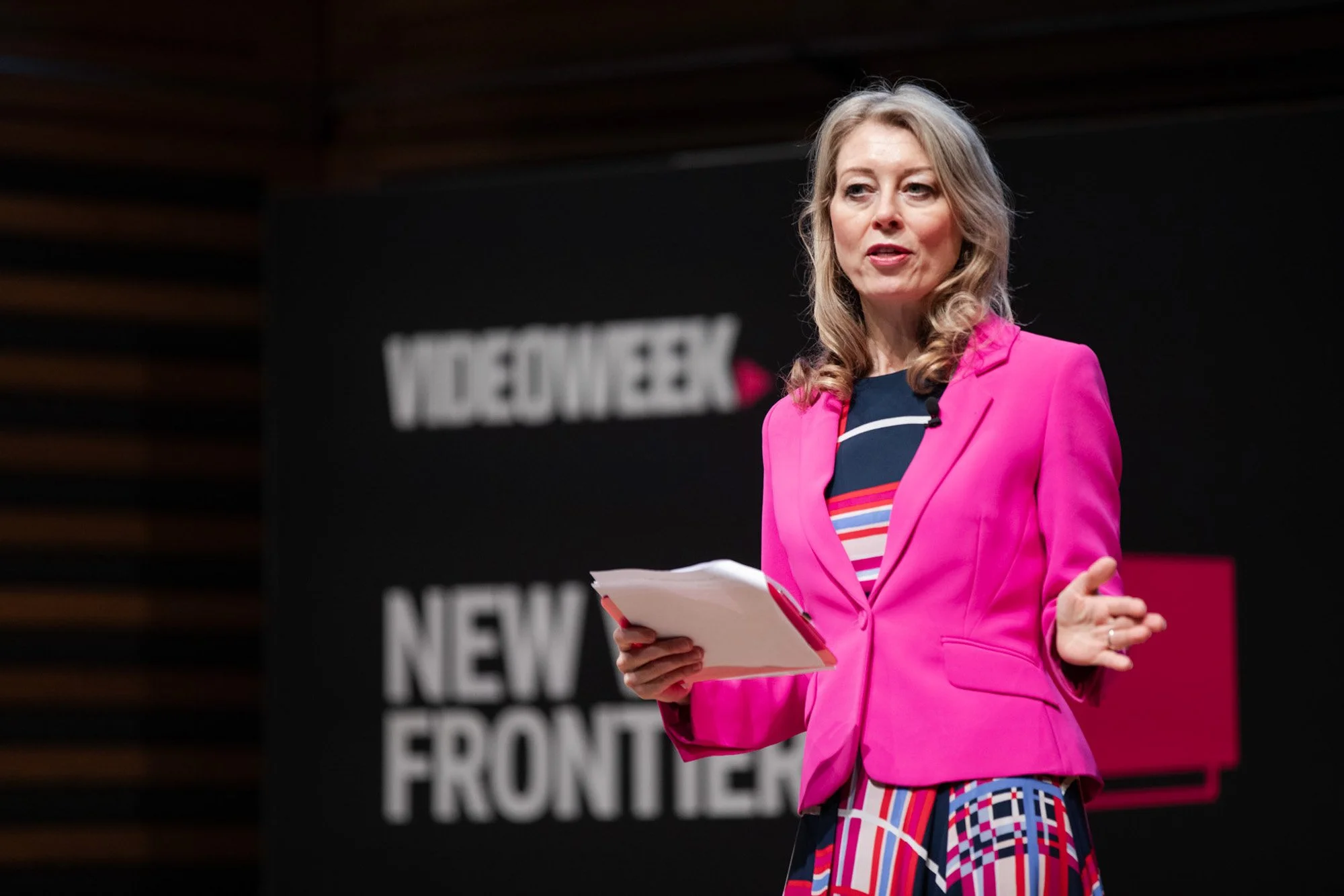 A woman in a pink jacket speaking on the New Video Frontiers stage.