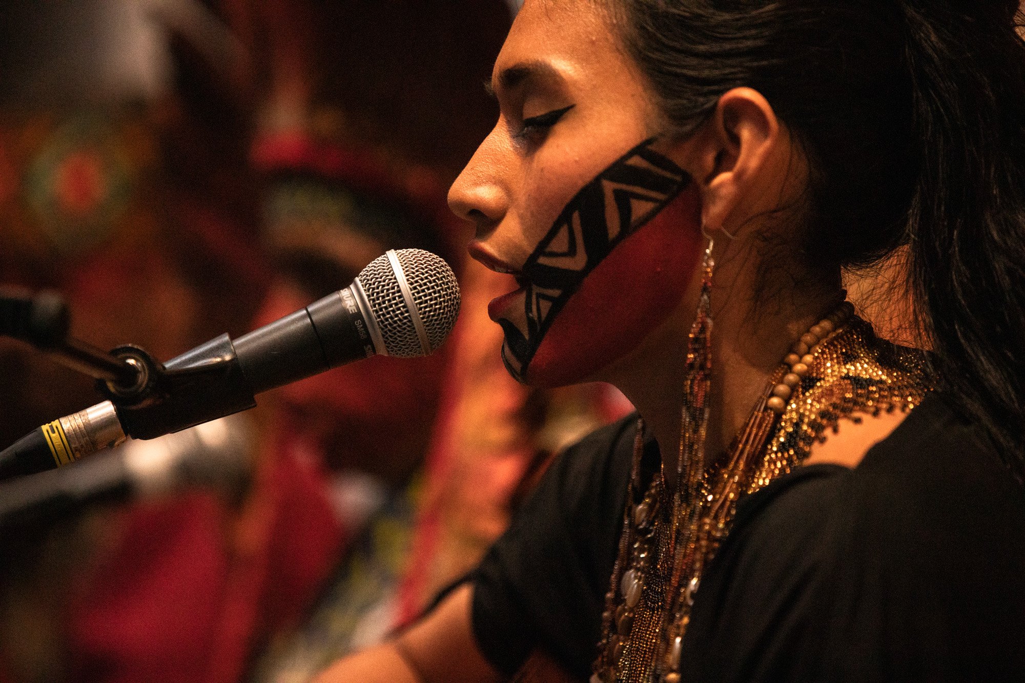 A singer with decorative face paint and gold jewellery performing into a microphone with eyes closed.