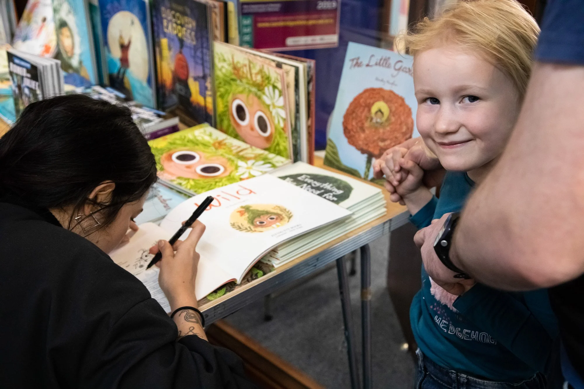 An illustrator drawing in an open picture book beside a smiling child at a book event.