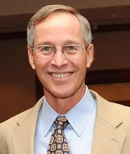 Headshot of a smiling man with glasses, wearing a beige suit, light blue shirt, and patterned tie, indoors.