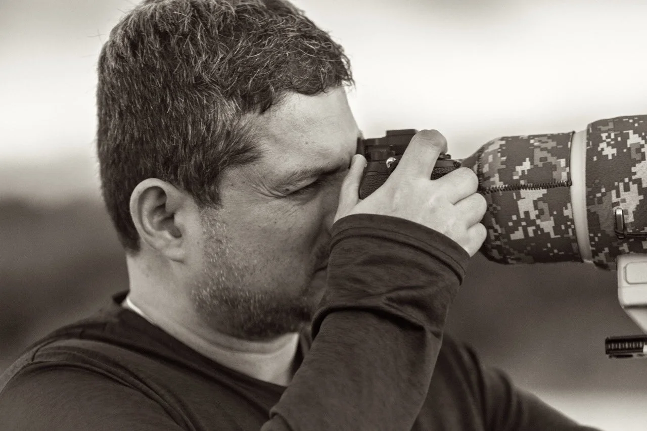 A man with short hair and a beard looking through the viewfinder of a camera, holding it with both hands and looking through the eyepiece.