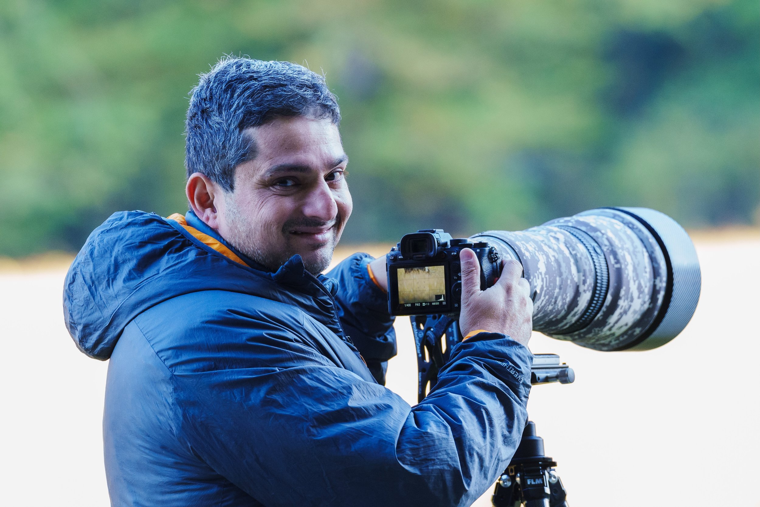 A man with dark hair and a beard, wearing a blue jacket, is smiling and looking at the camera while holding a professional camera with a large telephoto lens outdoors with a blurred green background.