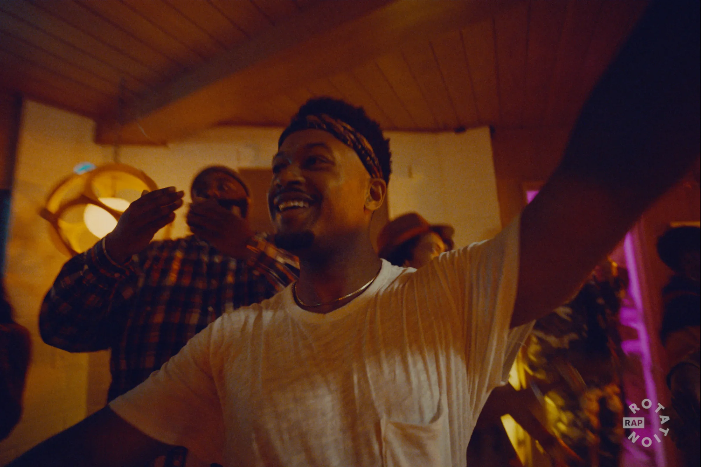 A Black man smiling at a social gathering, with friends in a warmly lit room with wooden ceiling, wearing a light-colored top and a patterned headband. Amazon Music commercial for Rap Rotation.