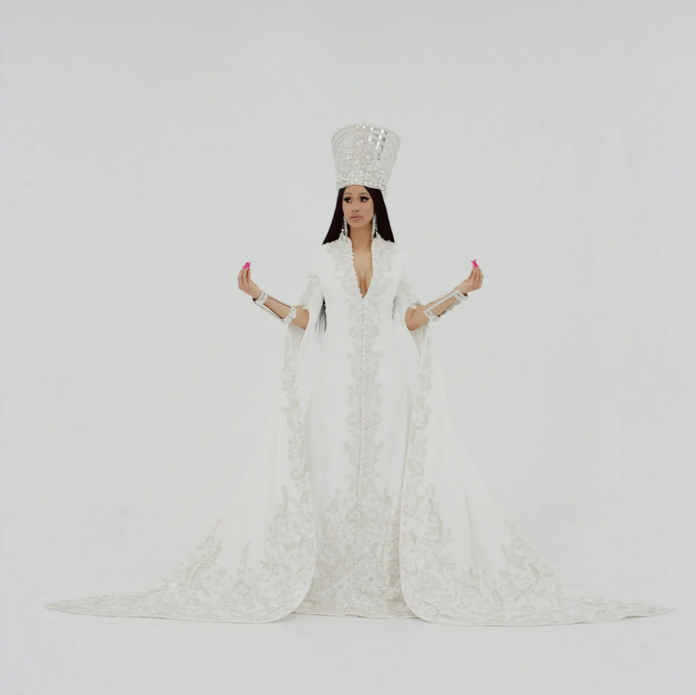 A woman wearing an elaborate white dress with lace, a large headpiece, and jewelry, standing against a plain white background.