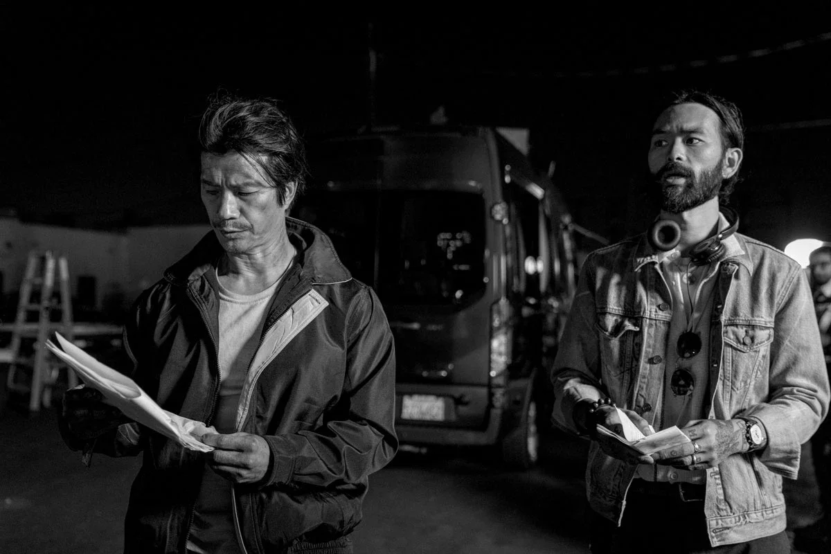Two men standing outdoors at night, one reading a script or document and the other holding papers. A bus is visible in the background. Director Sing J Lee, Actor Dustin Nguyen