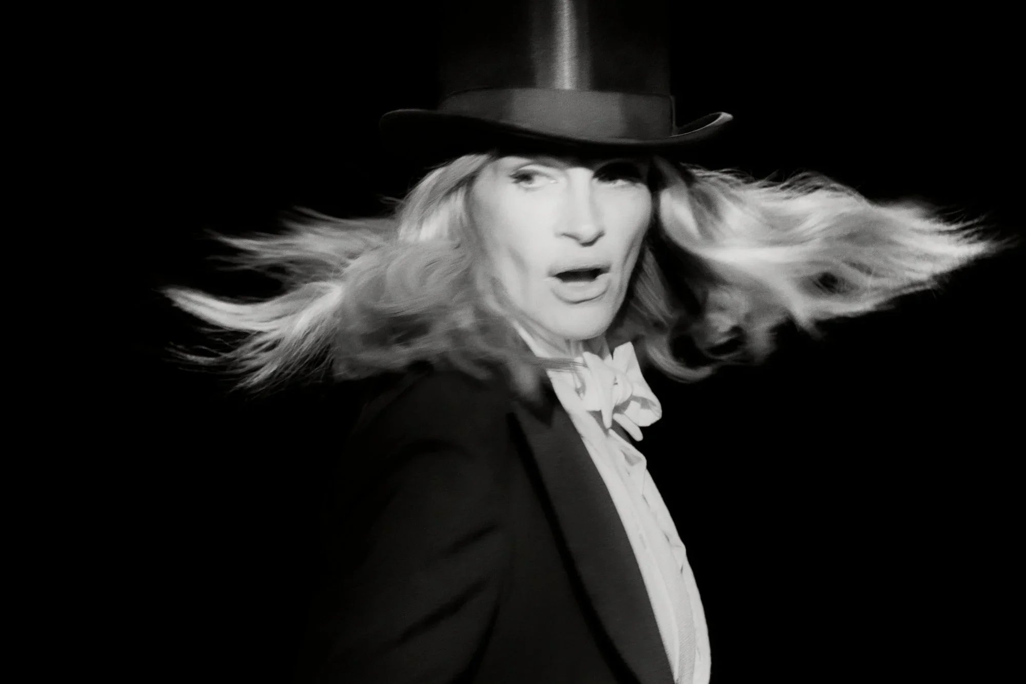 Black and white photo of a woman with long flowing hair, wearing a top hat and suit jacket, with an open-mouthed expression. Julia Roberts for New York Times Magazine photo by Philip Montgomery and choreography by Justin Peck.