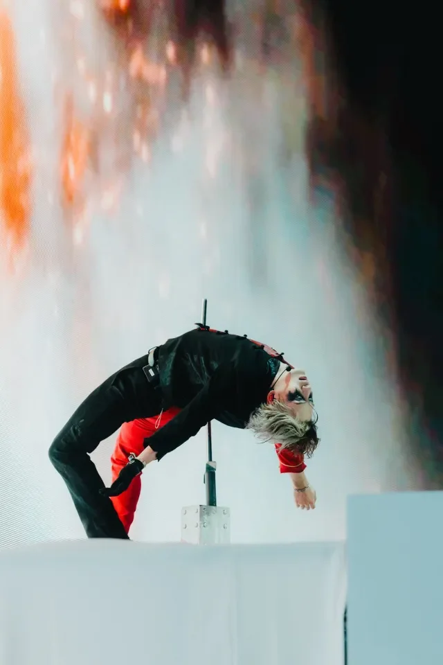 A performer on stage in a backbend position, with one hand touching the floor and the other hanging down, against a colorful backdrop.