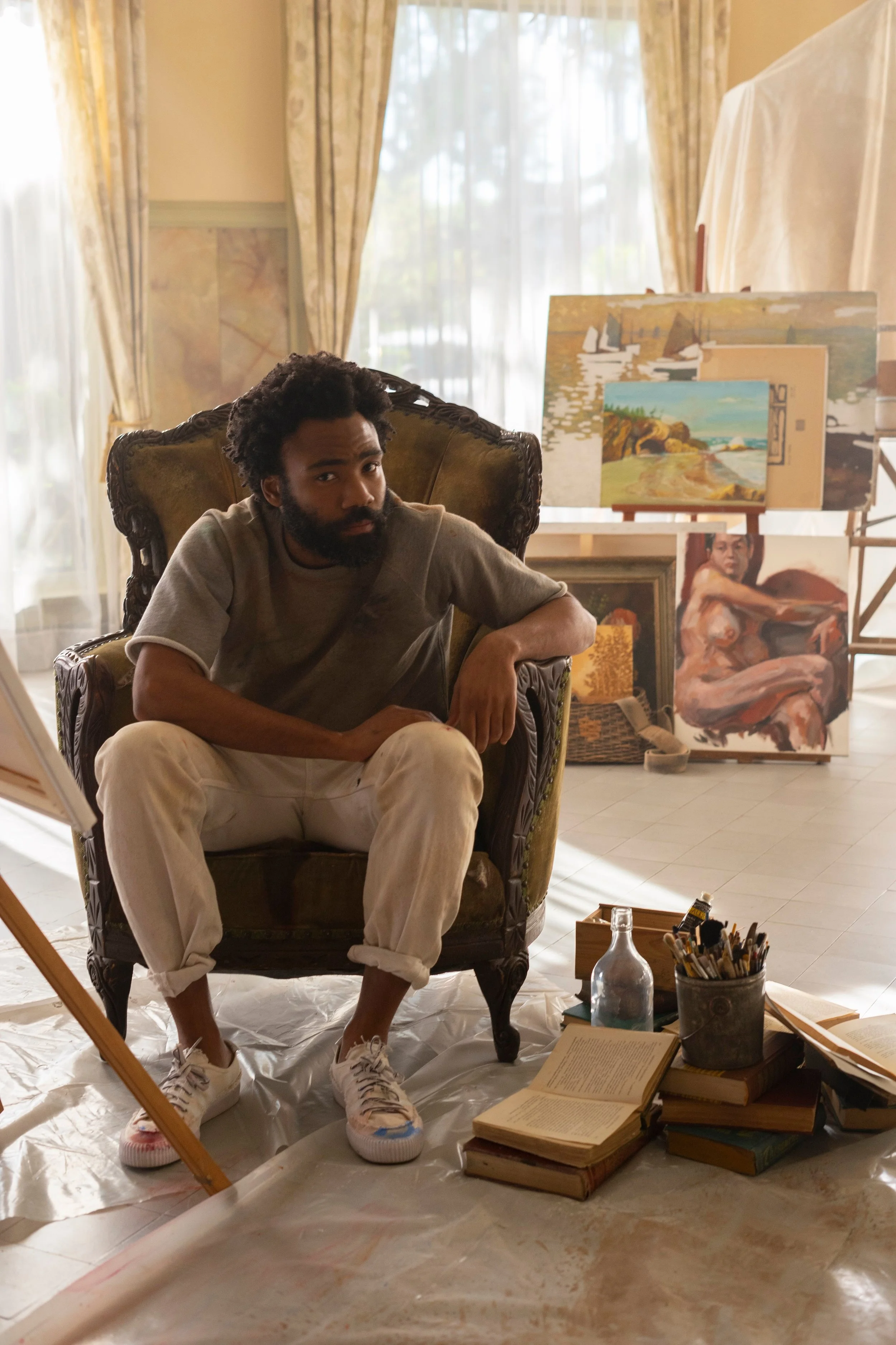 A man with a beard and curly hair sitting in an ornate armchair in an art studio, surrounded by paintings, books, and art supplies, with lots of natural light.