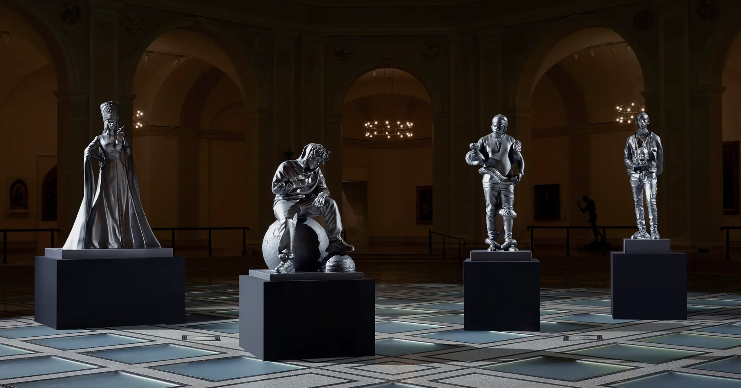 Four metallic sculptures of diverse individuals displayed on black pedestals inside a grand hall with high arched ceilings and chandeliers. Spotify Pantheon for Rapcaviar Brooklyn Museum.
