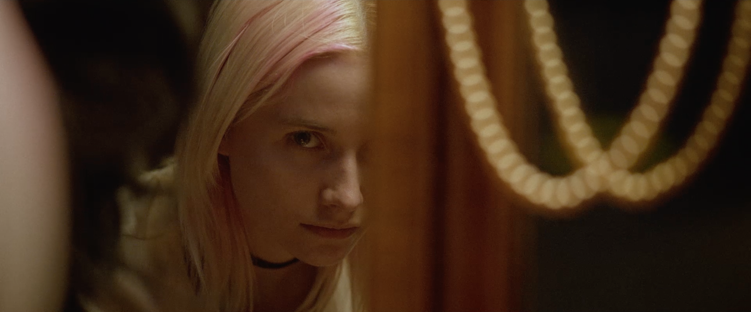 A woman with blonde hair peeking around the corner in a dimly lit setting, with a blurred string of warm-colored lights in the background.