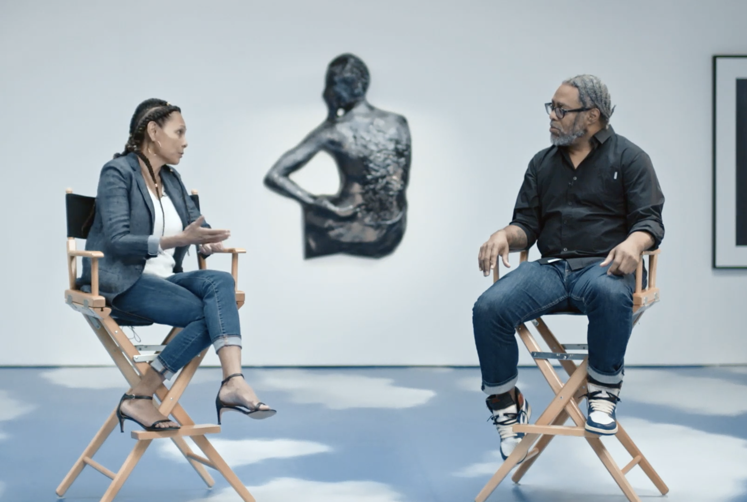 Two people are seated in director's chairs, engaged in conversation, with a sculpture of a torso in the background.