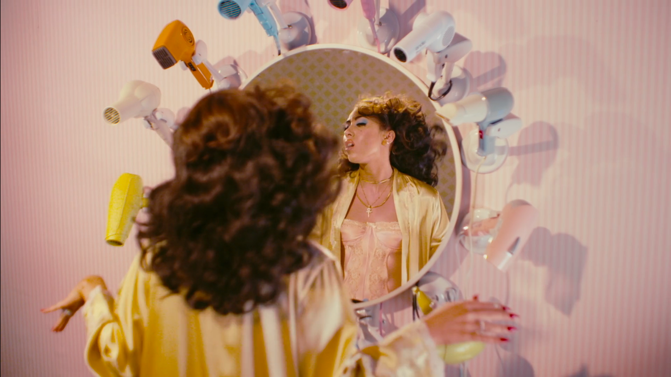 A woman with curly hair and blue eyeshadow looks at her reflection in a mirror, surrounded by a circular collection of hairdryers attached to a pink-striped wall.