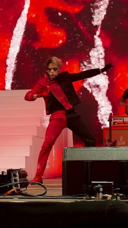 Performer on stage in a red and black suit, dancing with one arm extended, in front of a vibrant red and black digital background.