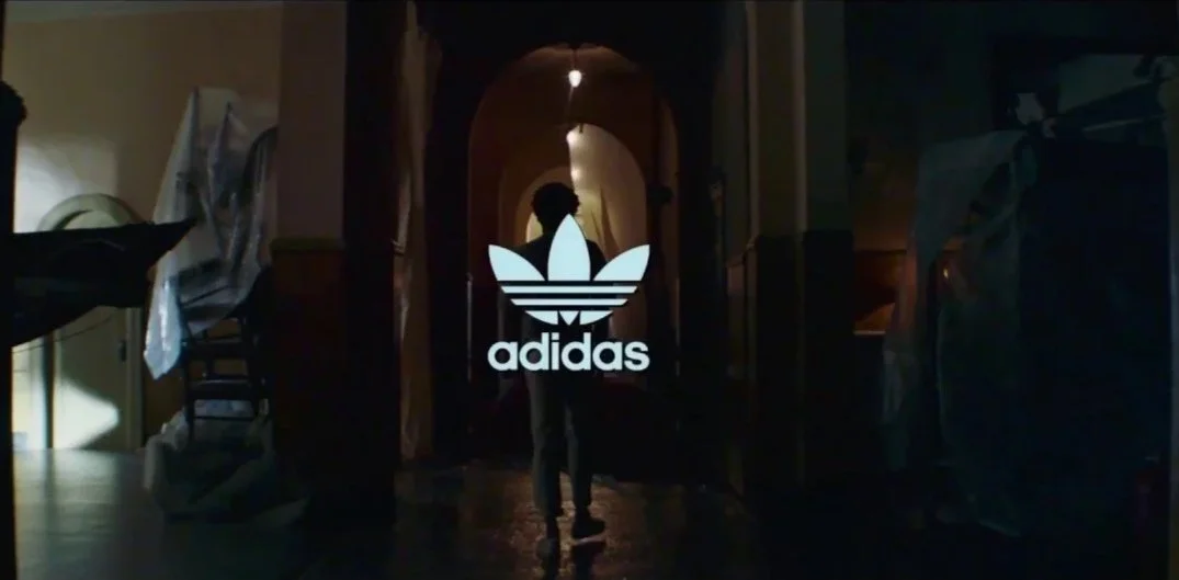 A person walking through a dimly lit corridor with a large Adidas logo on a glass door or window at the end.