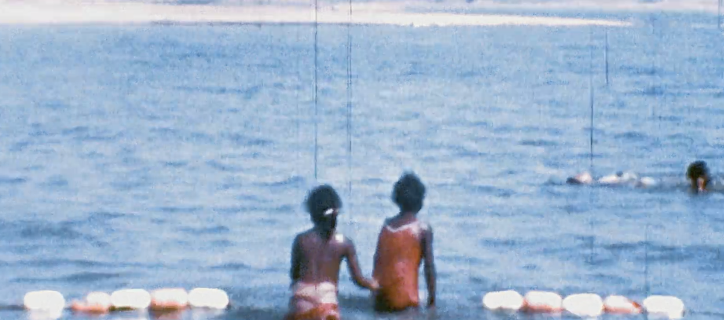 Two children standing side by side at the edge of a swimming pool, with other children swimming in the water and a swimmer in the pool.