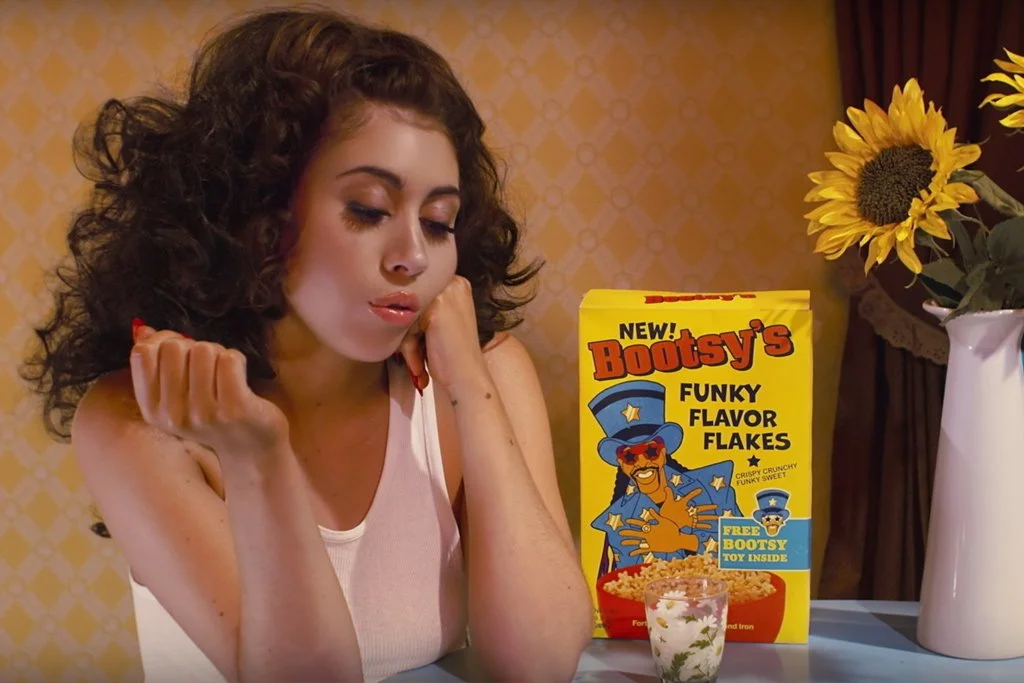 A woman with curly brown hair sitting at a table with a box of Bootsy's Funky Flavor Flakes cereal, a cup of milk, and a vase of sunflowers. She appears to be bored or upset.