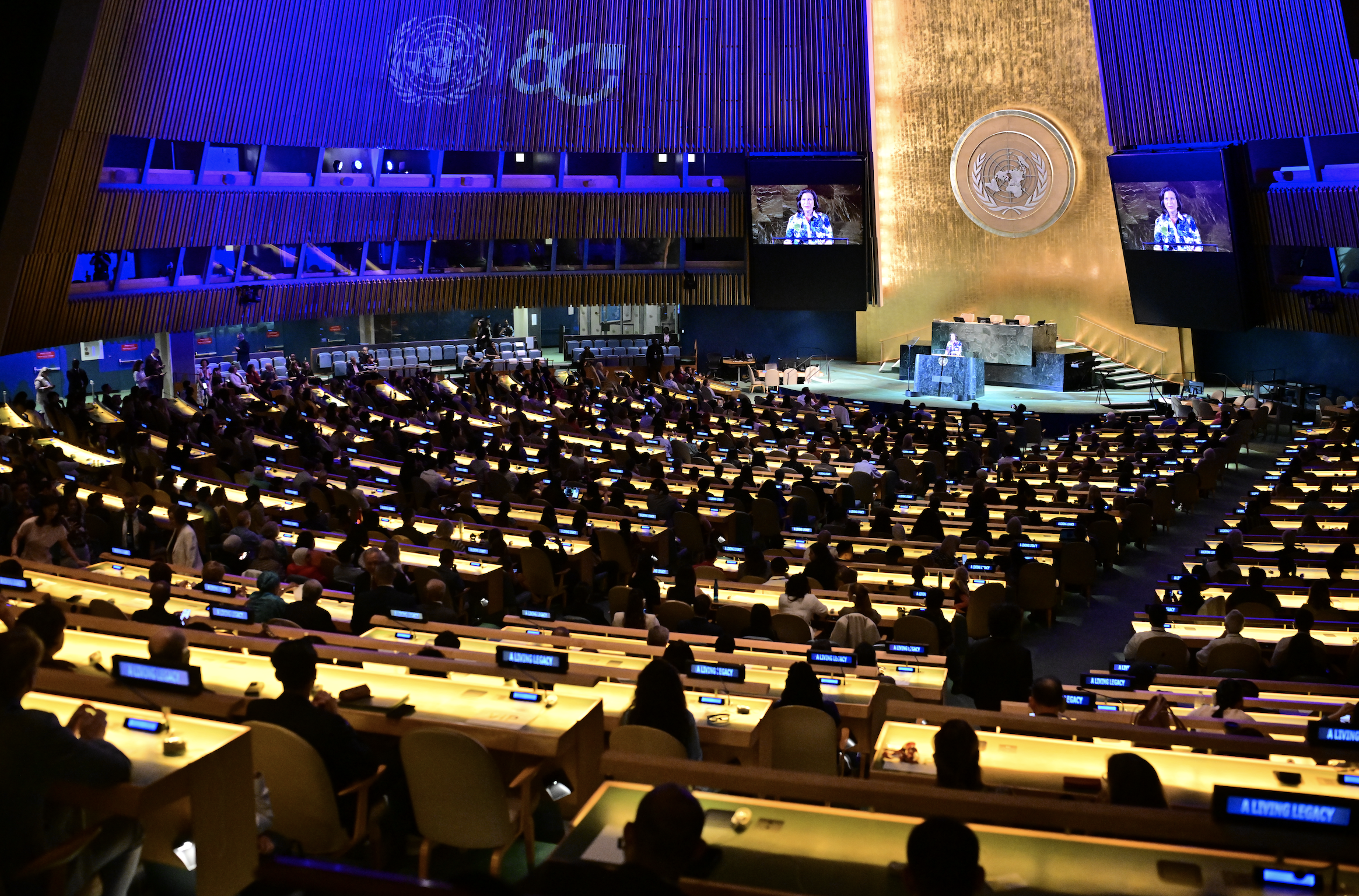 The United Nations General Assembly hall with people seated and a speaker at the podium, screens displaying the speaker, and the UN emblem on the wall.
