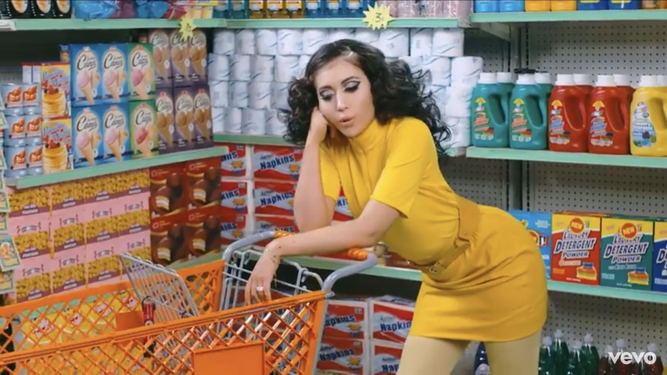 A woman with dark, curly hair and painted eyes in a yellow dress leaning on a grocery cart in a supermarket aisle, surrounded by shelves stocked with laundry detergent and cleaning supplies.