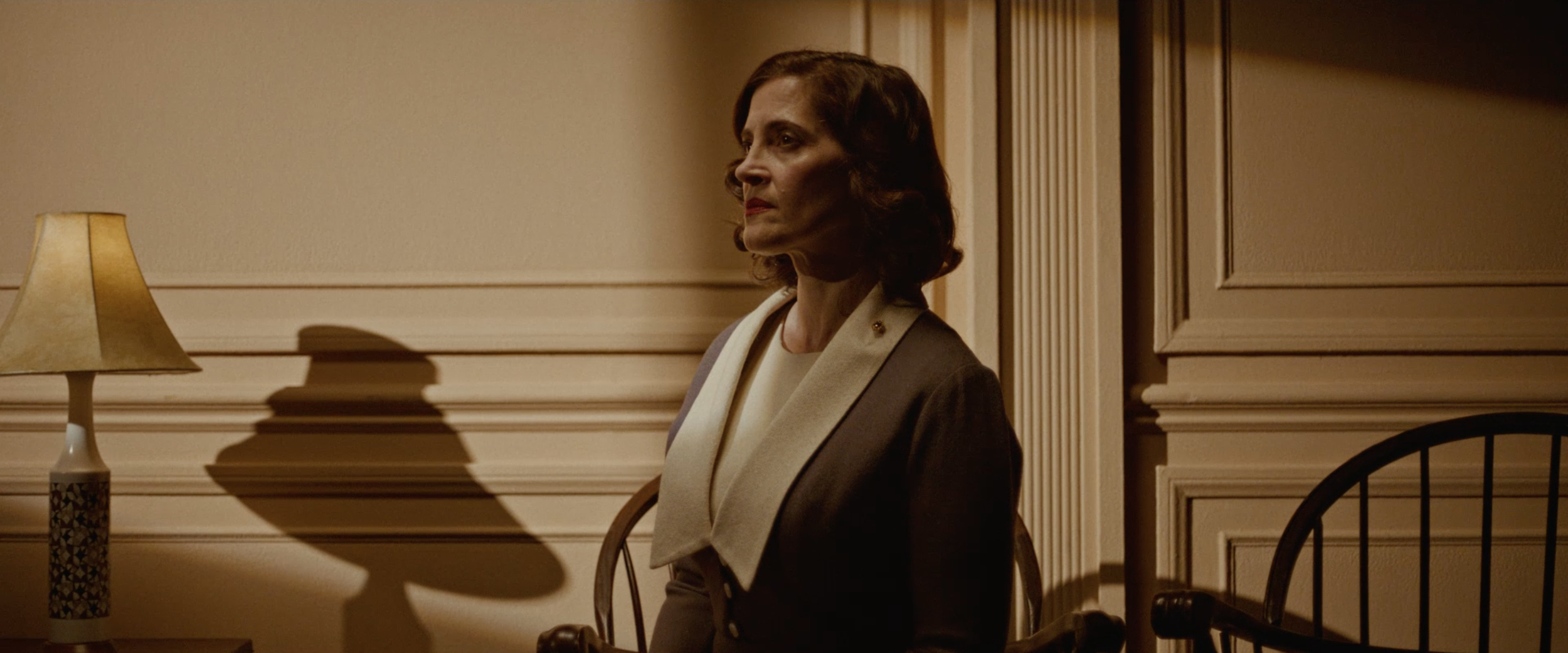 A woman with shoulder-length brown hair stands by a wall, looking to the left. She is wearing a grey blazer with white lapels over a white top. There is a beige lamp on a table to her left casting a shadow on the wall.