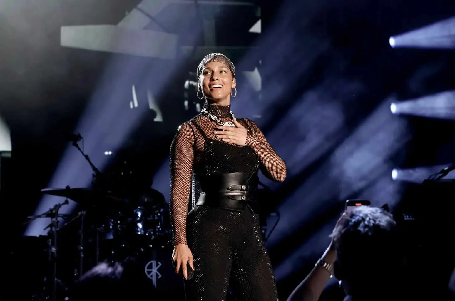 A woman singing or speaking on stage, wearing a glittery black outfit, jewelry, and a headpiece, with stage lights and musical instruments in the background. Alicia Keys for 2022 KEYS World Tour Live Nation.