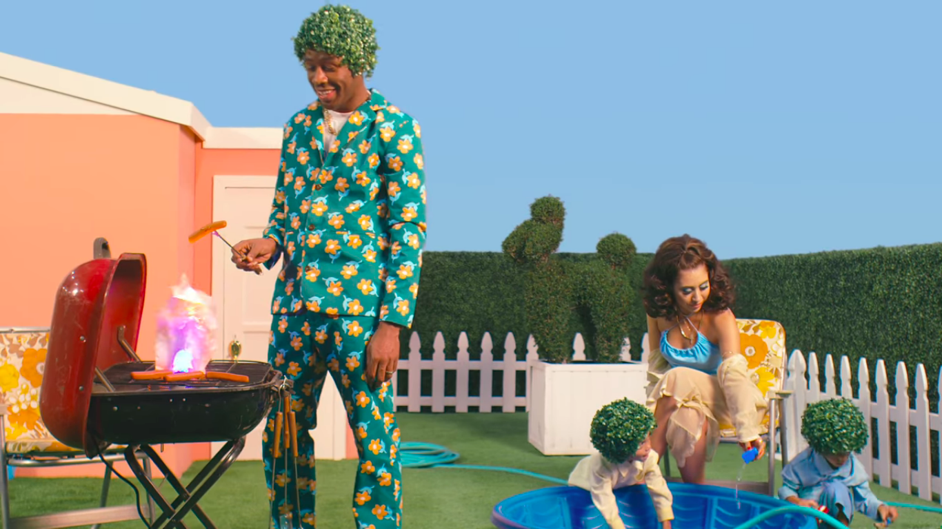 A man in a colorful floral suit with an afro-shaped tree wig is lighting a barbecue grill, which is producing flames. A woman sitting nearby is tending to two young children, who are playing with water in a small blue pool, with a white picket fence and shrubbery in the background.