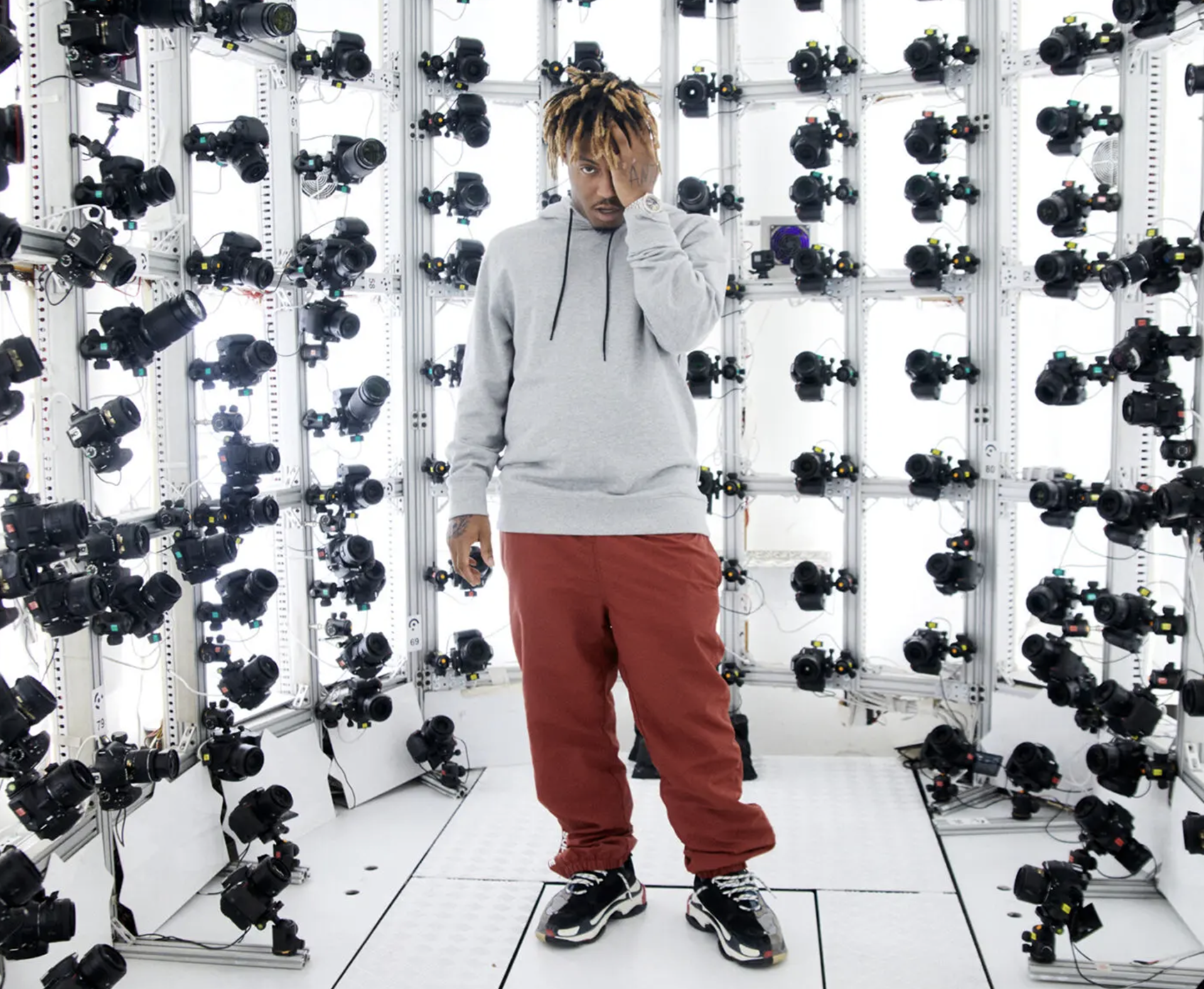 A man with blond dreadlocks and casual clothing standing on a platform among numerous cameras mounted on a wall in a white, high-tech room.