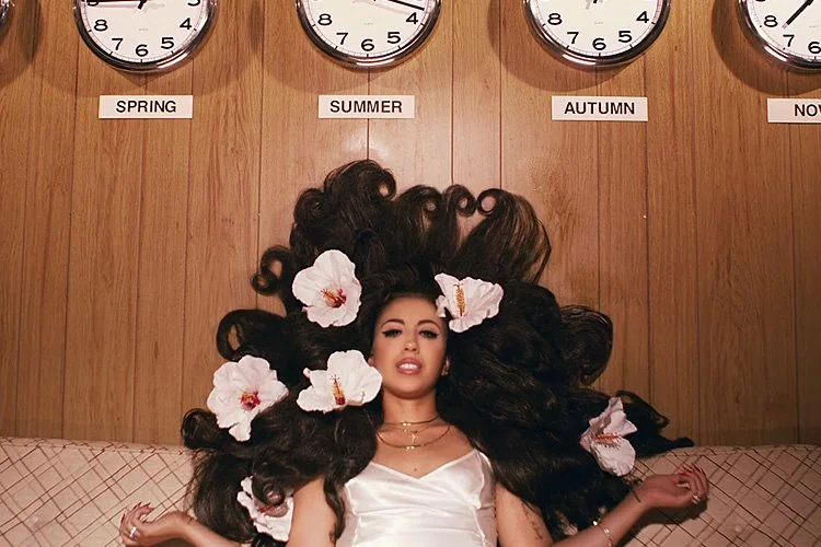 A woman with large, curly dark hair decorated with white hibiscus flowers, lying on a bed with a wooden wall background featuring four clocks labeled SPRING, SUMMER, AUTUMN, and WINTER.